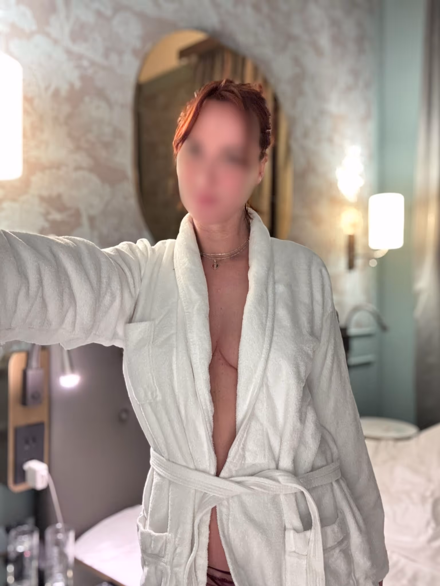 Eine Person steht in einem weißen Bademantel in einem Innenraum und hält einen Arm seitlich ausgestreckt; im Hintergrund sind Bett, Lampe und Wandmuster zu sehen.