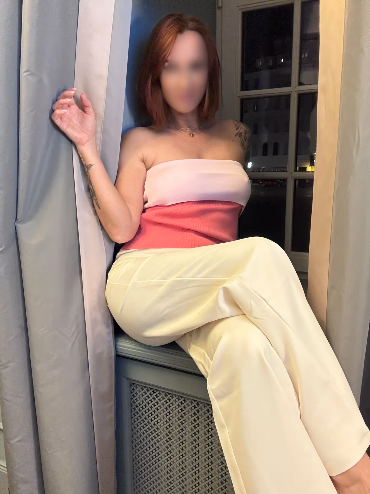 Eine Person sitzt seitlich auf einer Fensterbank oder Heizung vor einem Fenster und trägt ein trägerloses Oberteil in Rosa und Weiß sowie eine helle Hose.