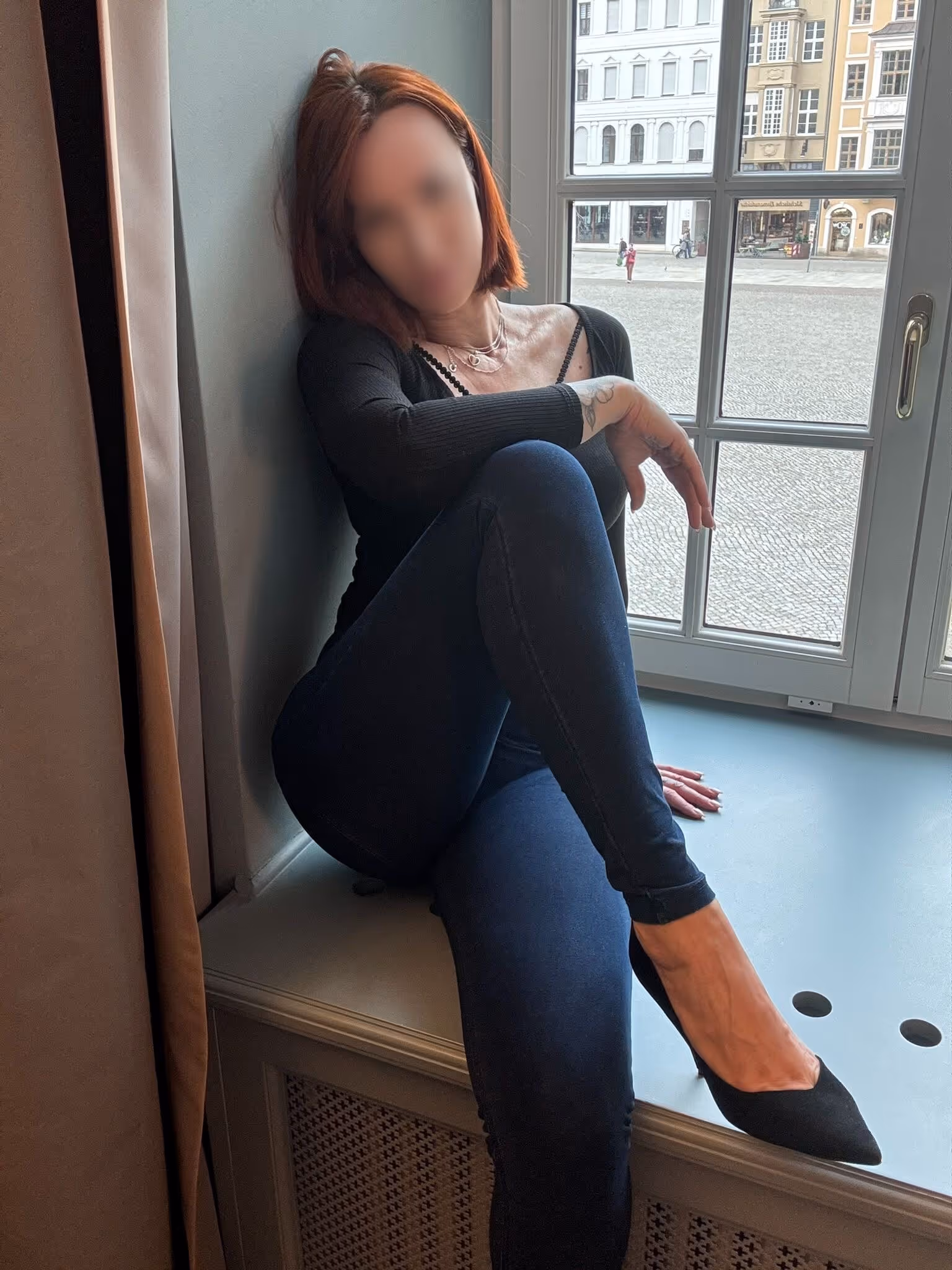 Emily sitzt seitlich an einem Fenster und trägt ein schwarzes langärmliges Oberteil mit dunkler Jeans; die Aufnahme zeigt eine sitzende Pose in einem Innenraum.