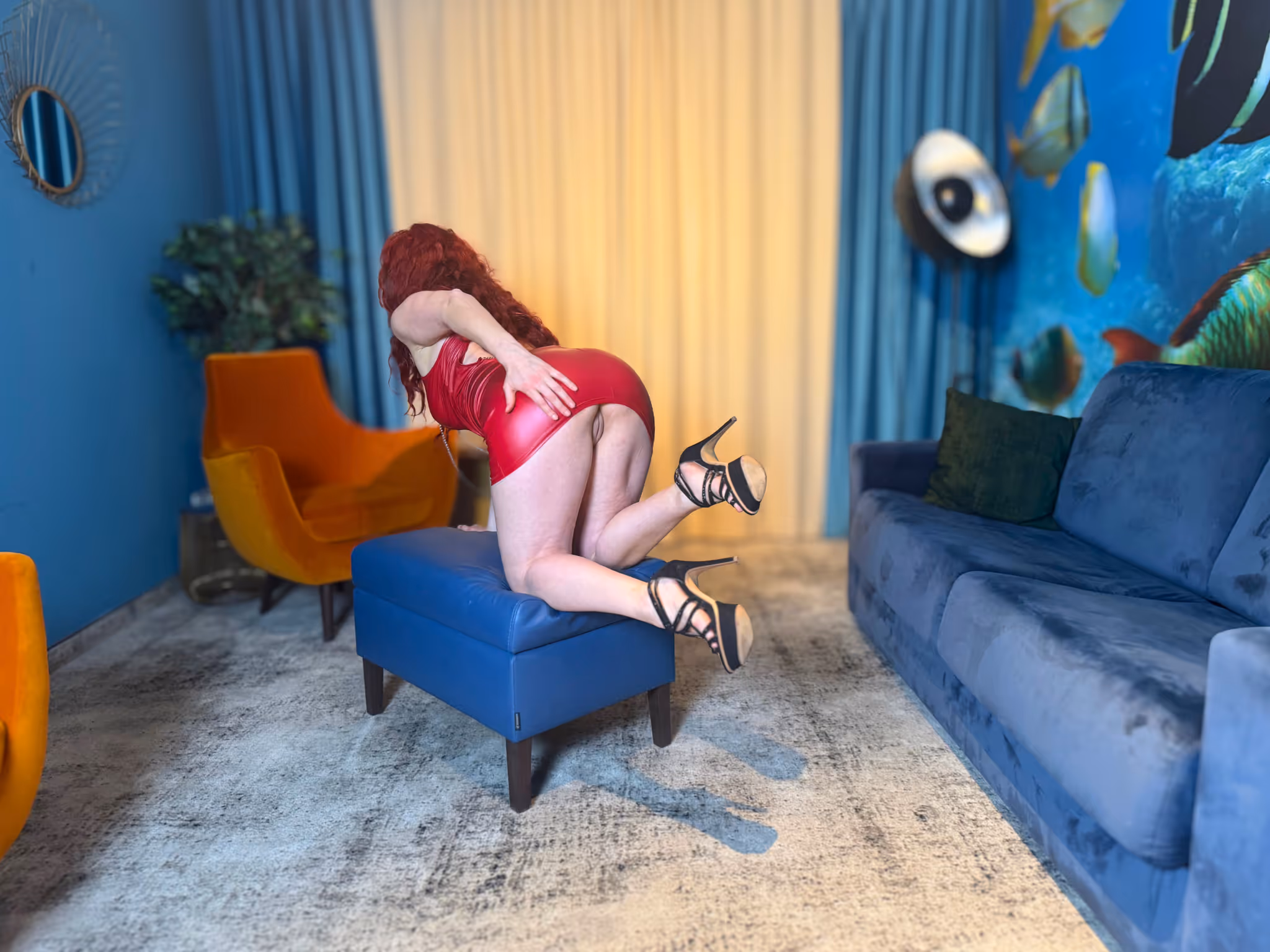 Escort Romy Bondage in Dresden kniet auf dem blauen Samt Sessel in knallrotem Bondage Bikini mit Riemen und String. Lange rote Locken fallen über den Rücken schwarze High Heels Hände hinter dem Kopf gefesselt verführerische und devote Bondage Pose Aquarium Ambiente Dresden.
